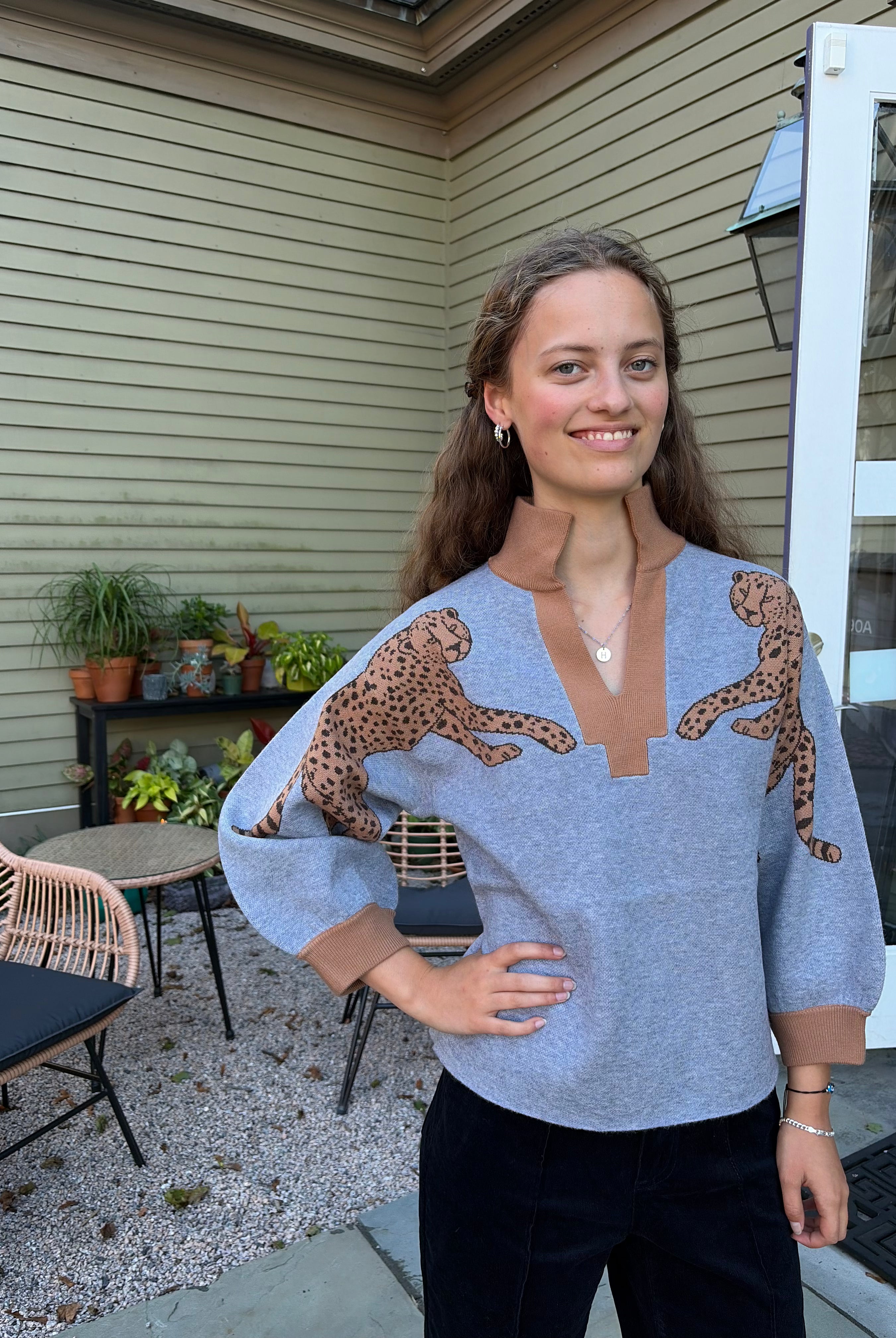 A woman wearing a light blue knit sweater with brown cheetah prints on the shoulders and brown ribbed trim, standing on a patio in front of a house with beige siding.