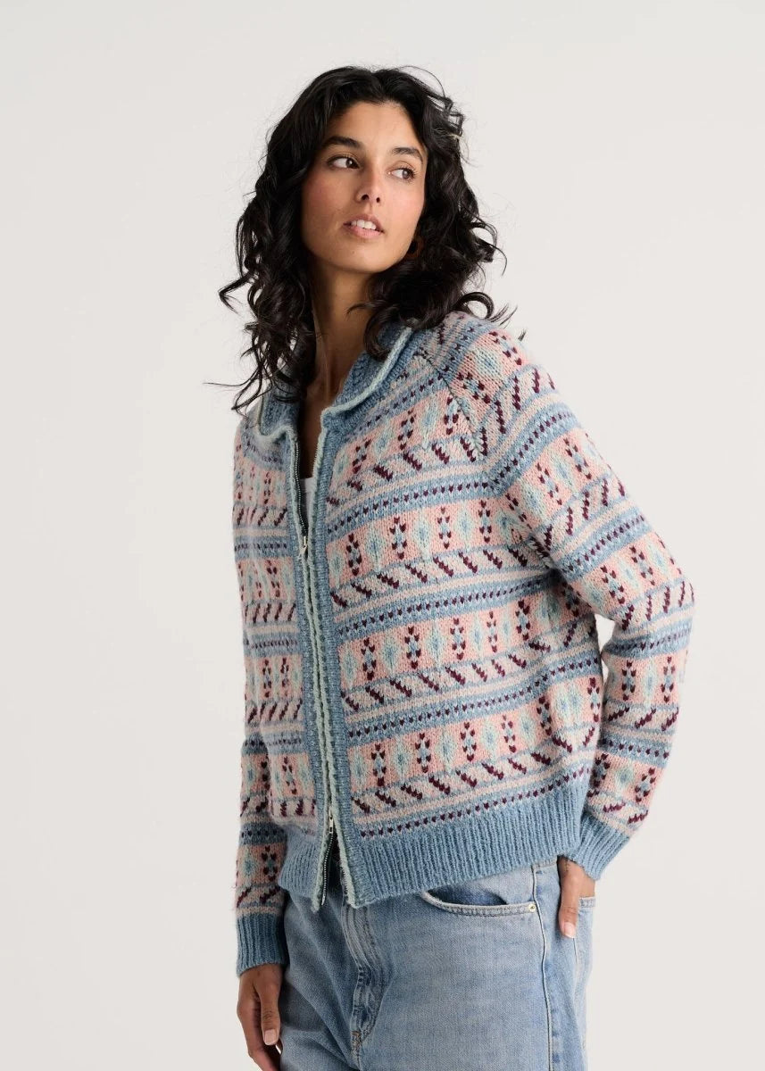 A woman wearing a light blue knit zip-up cardigan with pink and white geometric patterns, paired with light-wash blue jeans, against a plain light gray background.