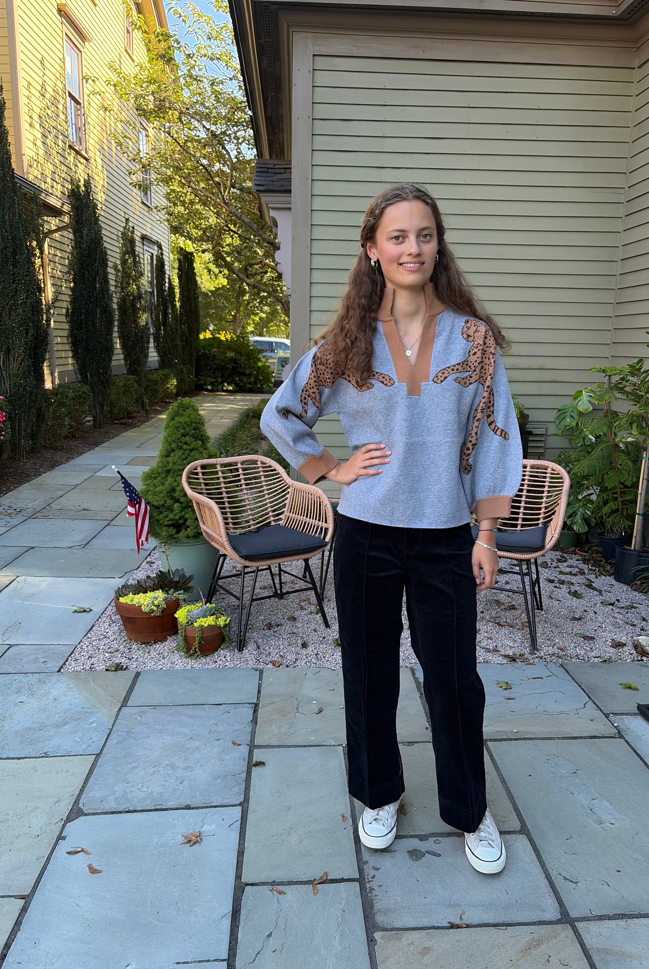A woman stands on a stone patio in front of a house, wearing a light blue knit sweater with brown and gold patterned shoulder details, black wide-leg pants, and white sneakers.