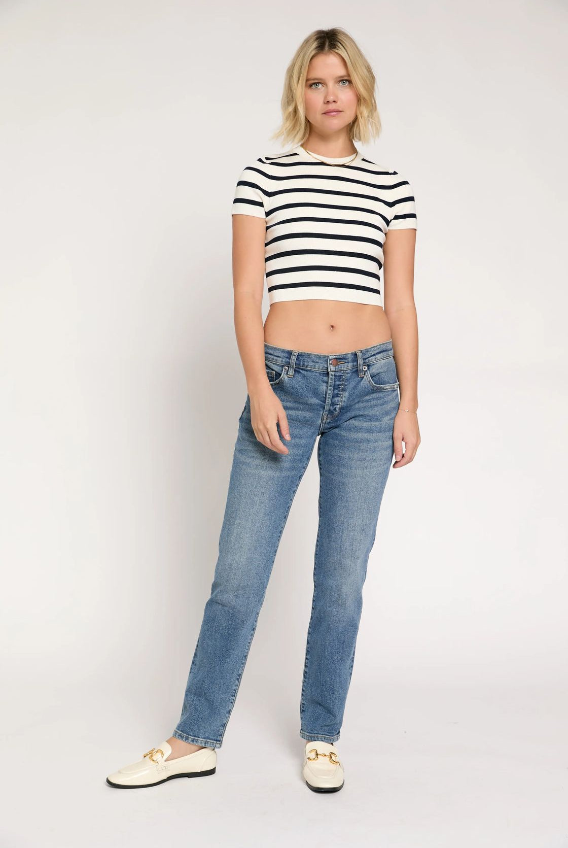 A woman modeling a black and white horizontally striped short-sleeve crop top with blue jeans and cream-colored loafers, standing against a plain light gray studio background.