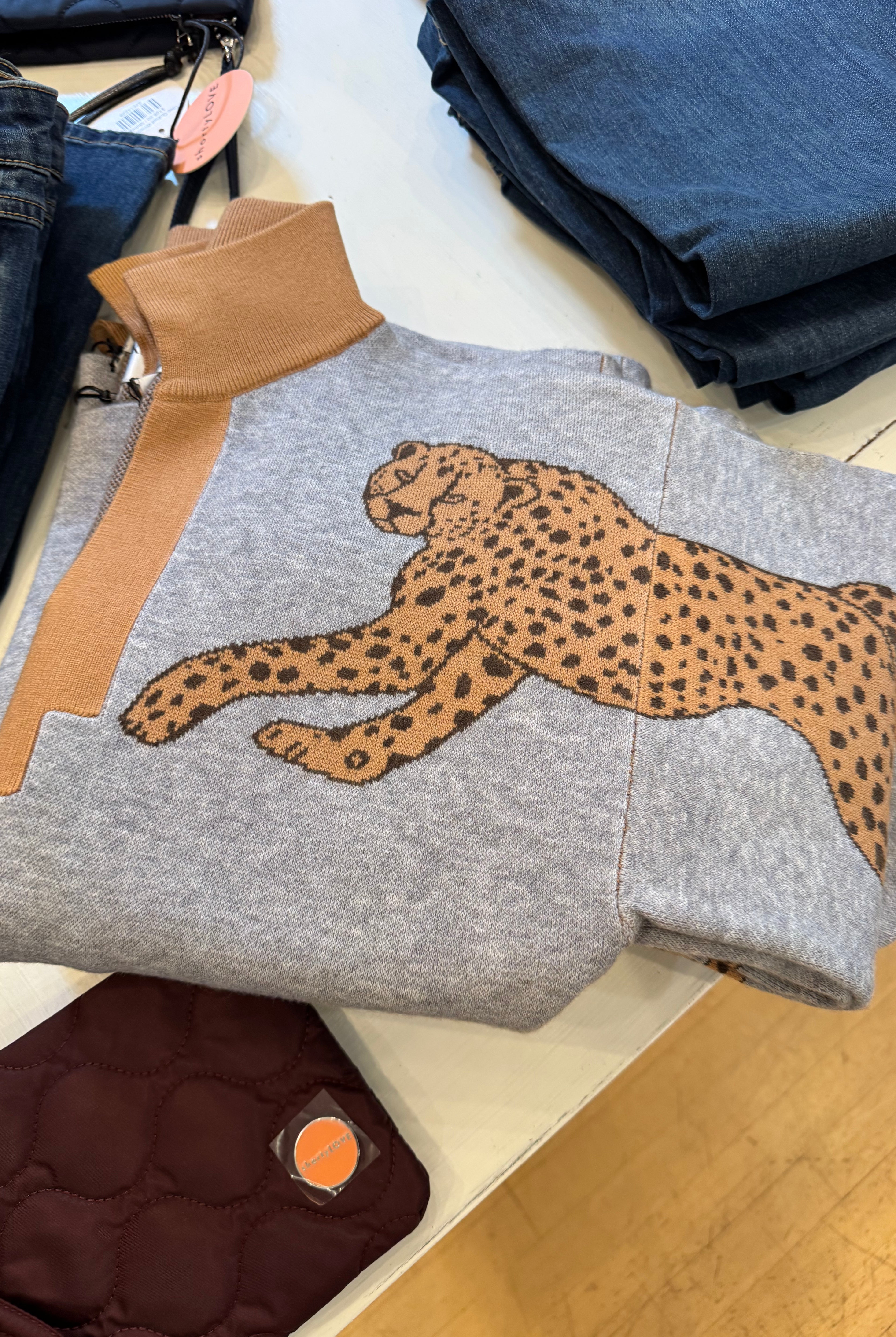 A folded heather gray sweatshirt with a large tan and black cheetah graphic across the chest, tan ribbed collar and cuffs, and a light wood-toned surface underneath. Other folded garments are partially visible around it.