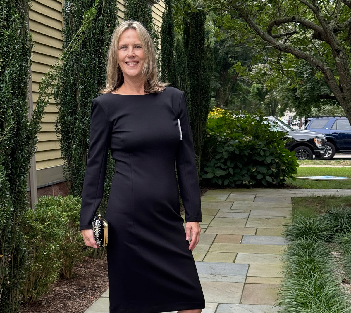 A woman stands on a stone walkway outdoors wearing a fitted black long-sleeve knee-length dress with structured shoulders, paired with black strappy heels and holding a small metallic clutch.