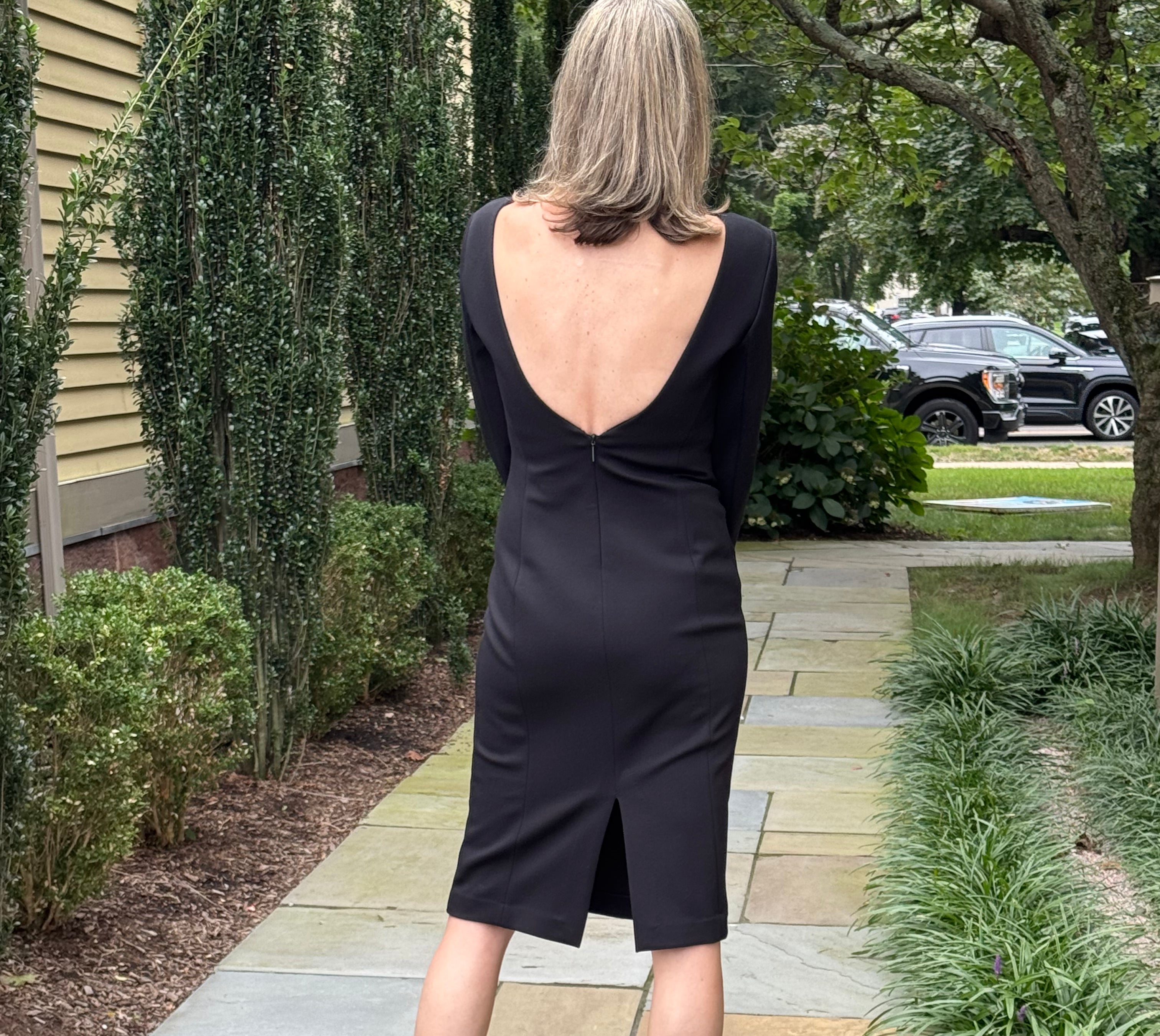 A woman stands on a stone walkway outdoors, photographed from behind, wearing a black long-sleeve dress with a deep open back and a small front slit.