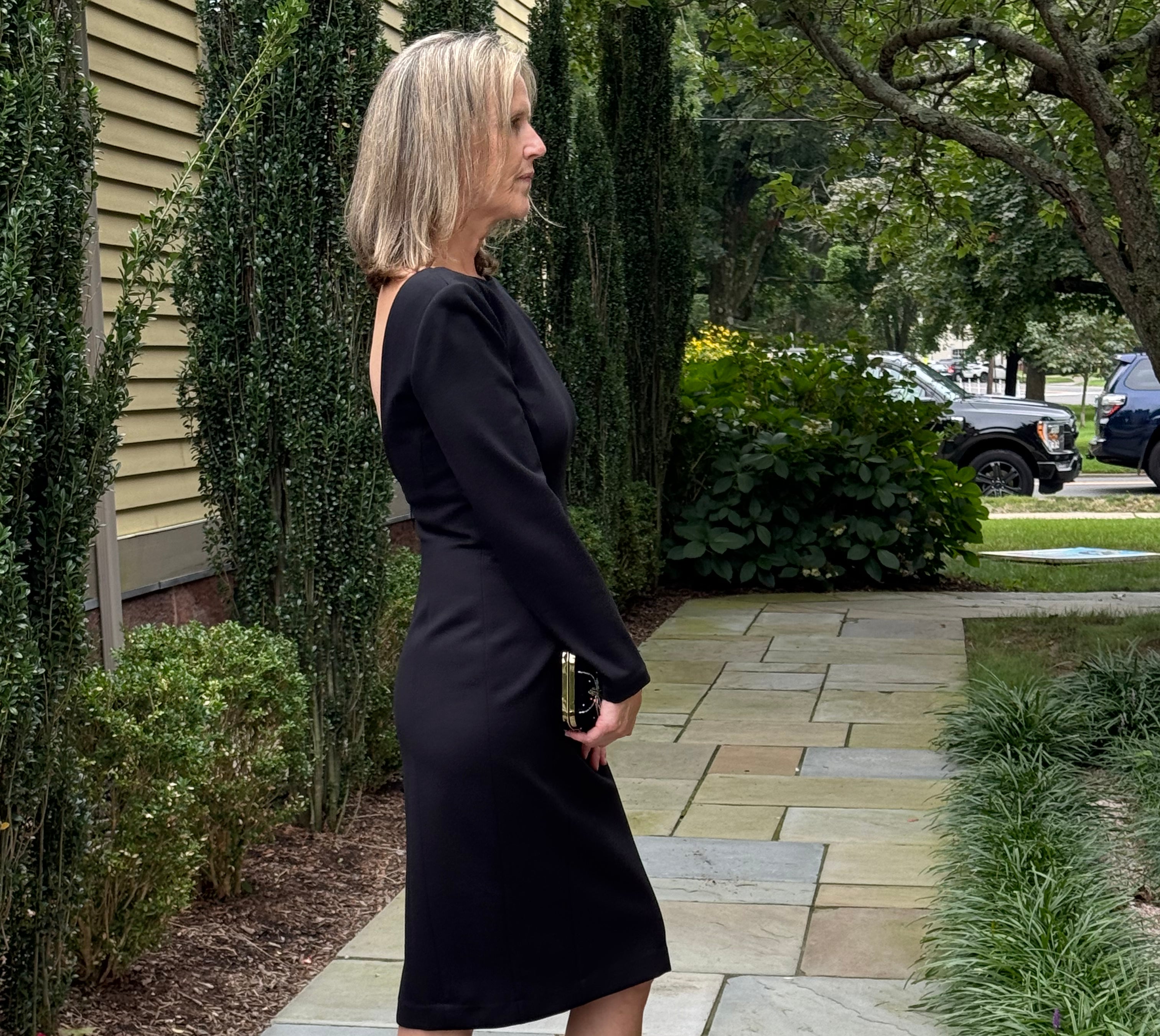 A woman stands on a stone walkway outdoors wearing a fitted black knee-length dress with long sleeves and black strappy heeled sandals, holding a small metallic clutch.