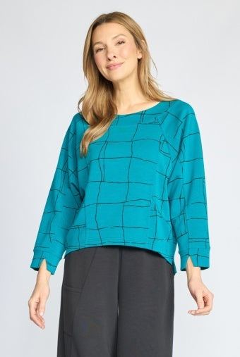 A woman modeling a teal long-sleeve blouse with a black geometric grid pattern, paired with dark gray wide-leg trousers, against a light gray studio background.