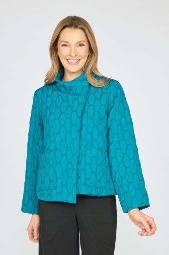 A smiling woman wearing a bright turquoise quilted jacket with a subtle geometric pattern, paired with black pants, against a light gray studio background.