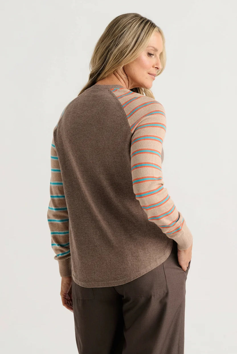 A woman shown from the back wearing a brown knit top with peach and blue striped raglan sleeves, paired with dark brown pants, against a light gray studio background.