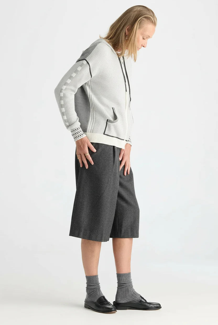 A model wearing a light gray and white knit zip-up hoodie with gray sleeve patches, dark gray knee-length shorts, gray socks, and black loafers, photographed against a plain light gray studio background.