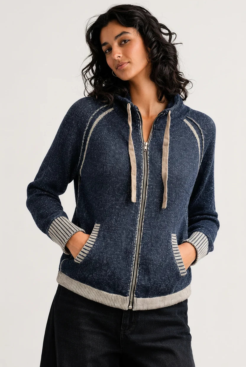 A woman wearing a dark blue zip-up hoodie with beige ribbed trim and drawstrings, paired with black pants, photographed against a light gray studio background.