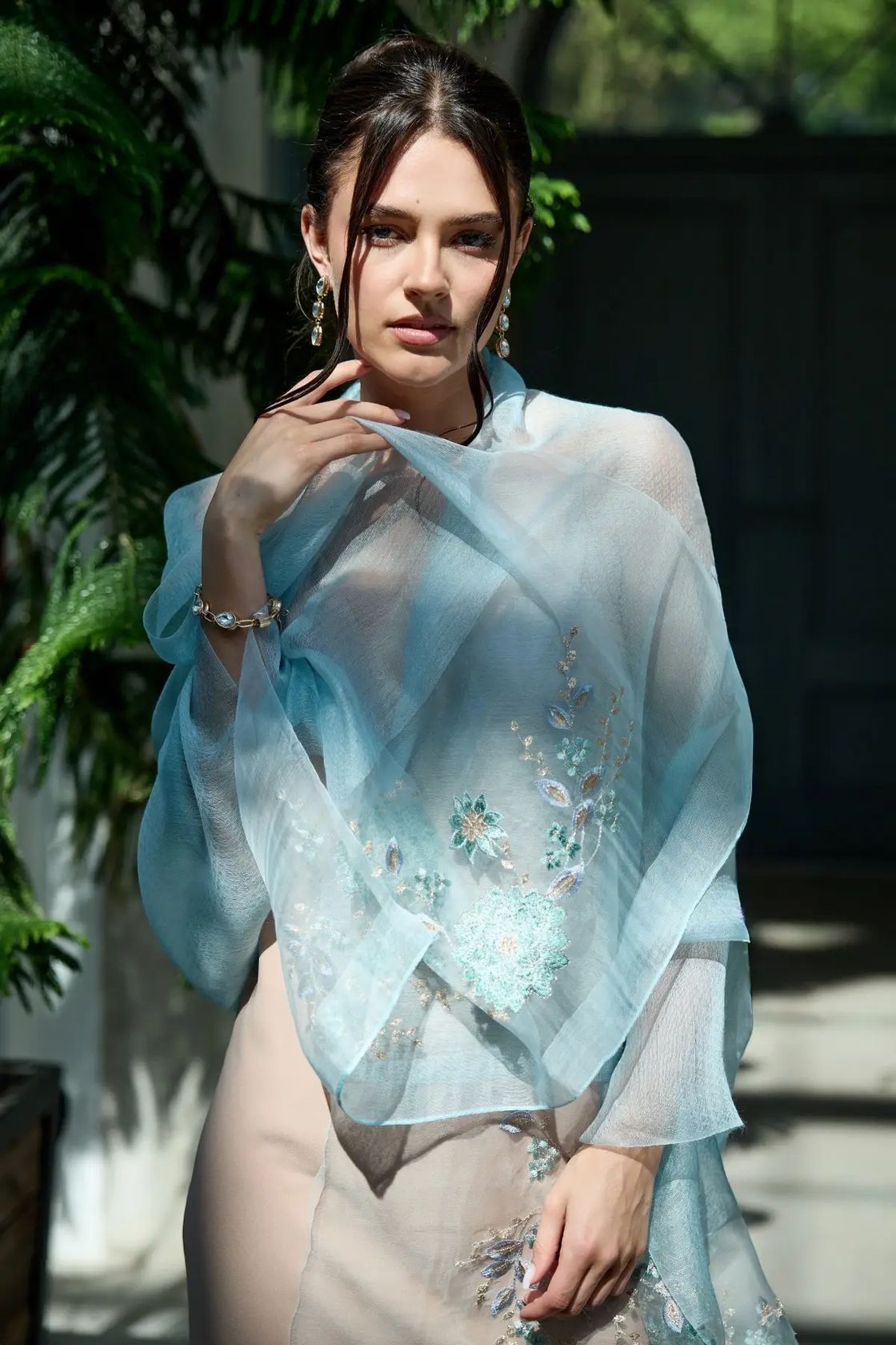 A woman wearing a sheer light blue embroidered shawl draped over her shoulders, paired with a beige dress, set against a background of green foliage and a dark wooden door.