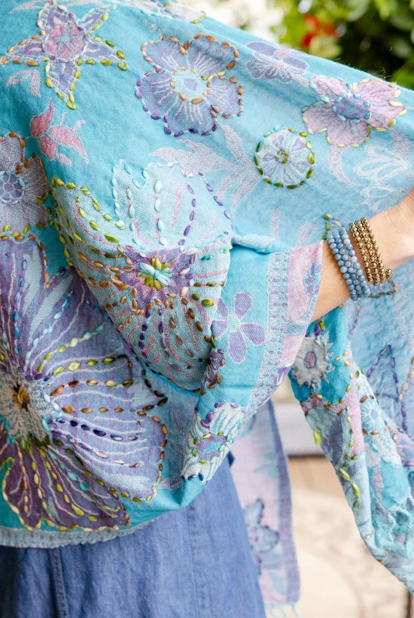 A light blue bohemian shawl with large purple and lavender floral embroidery, held up by a model wearing blue jeans and beaded bracelets.