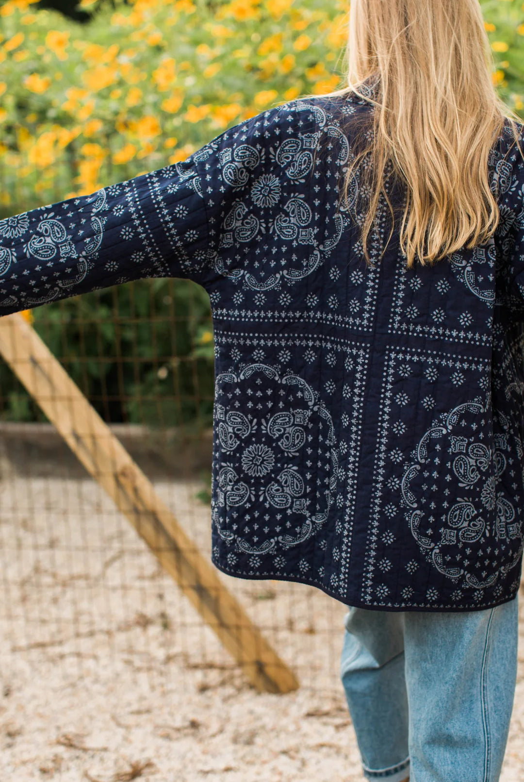 A woman viewed from behind wearing a navy blue bandana print cardigan over a light blue top and light wash jeans, standing outdoors in front of yellow flowers.