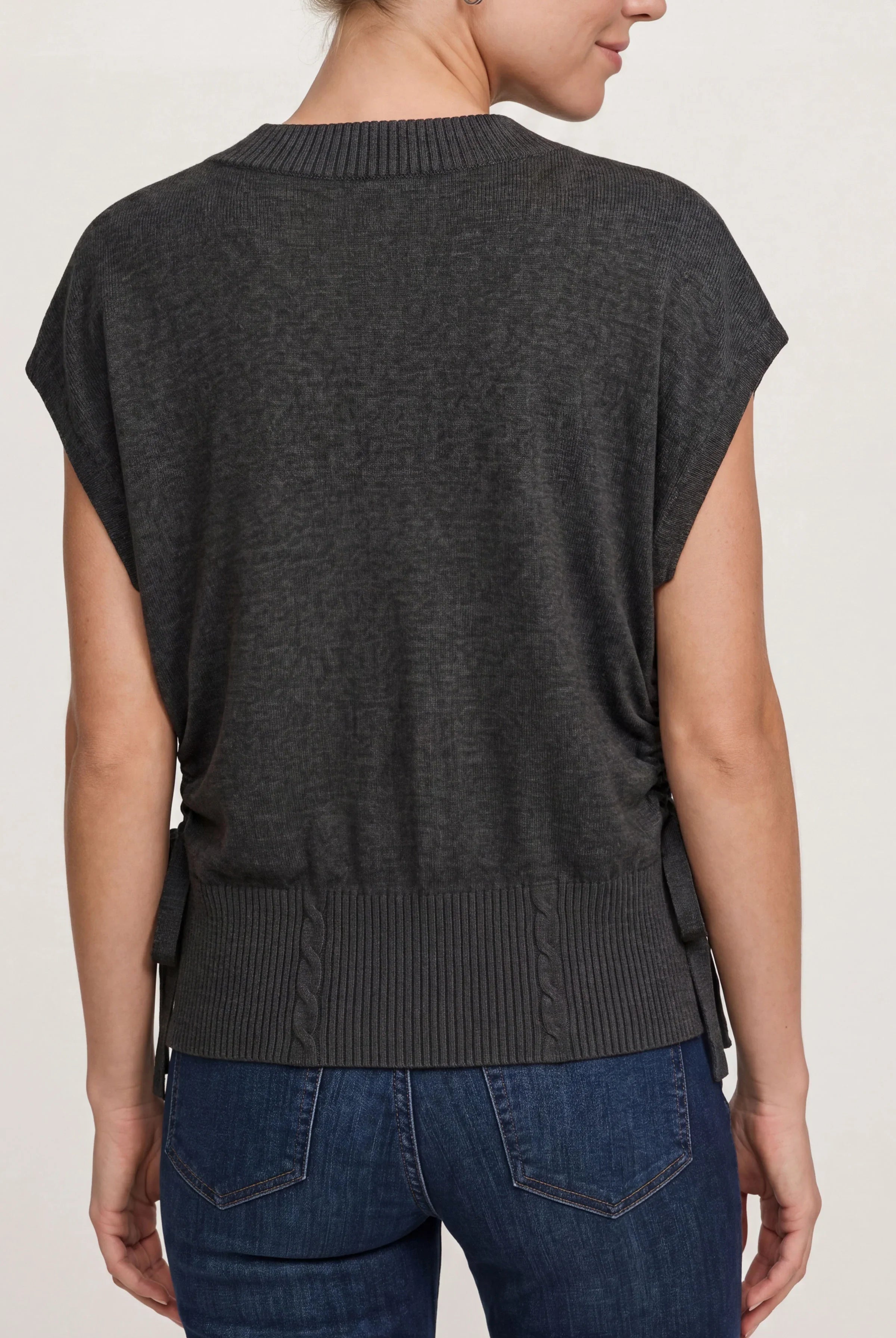 A woman shown from the back wearing a dark charcoal gray knit sweater with short dolman sleeves and a ribbed hem, paired with blue jeans against a light gray studio background.