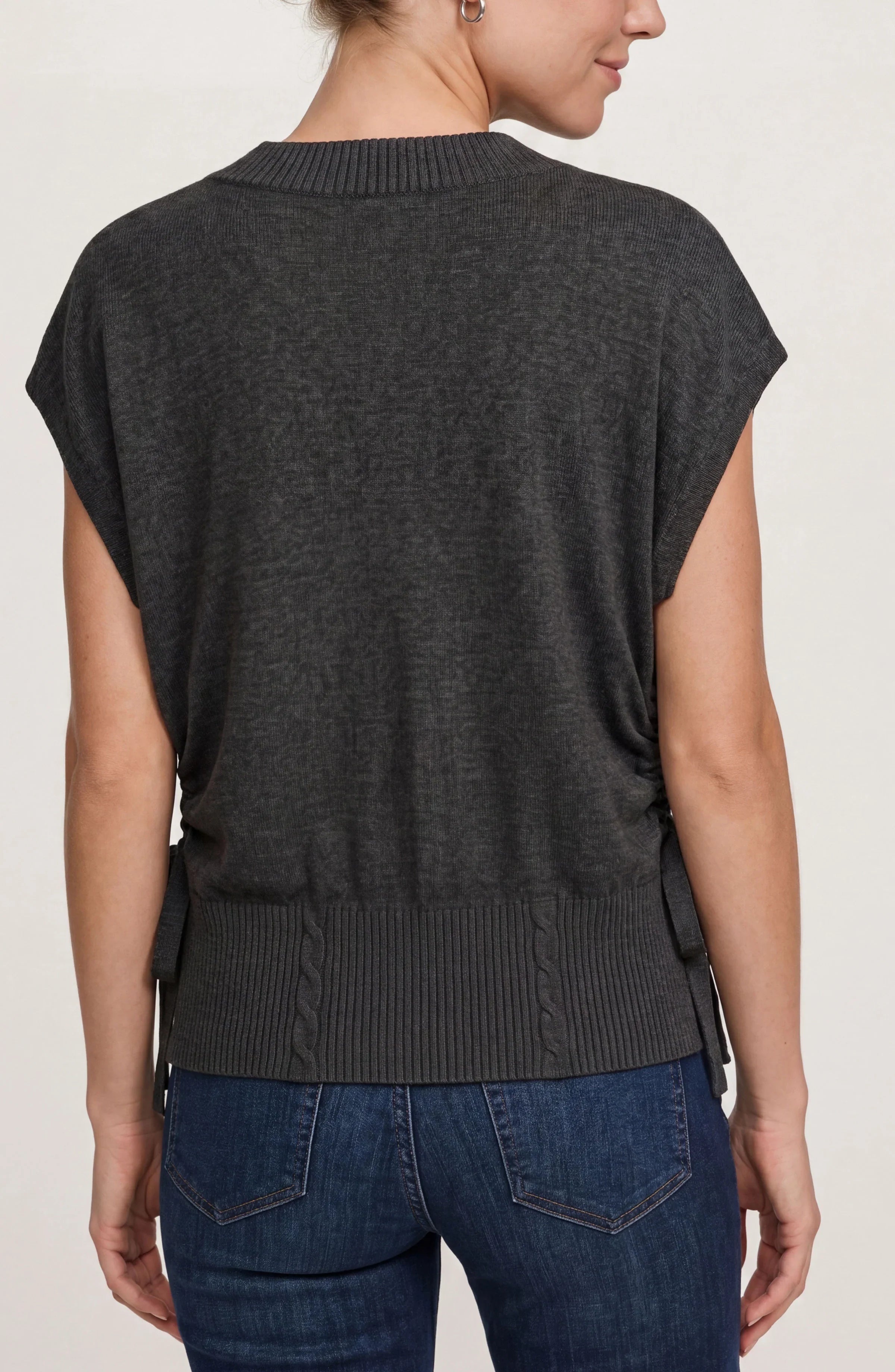 A woman shown from the back wearing a dark charcoal gray knit sweater with short dolman sleeves and a ribbed hem, paired with blue jeans against a light gray studio background.