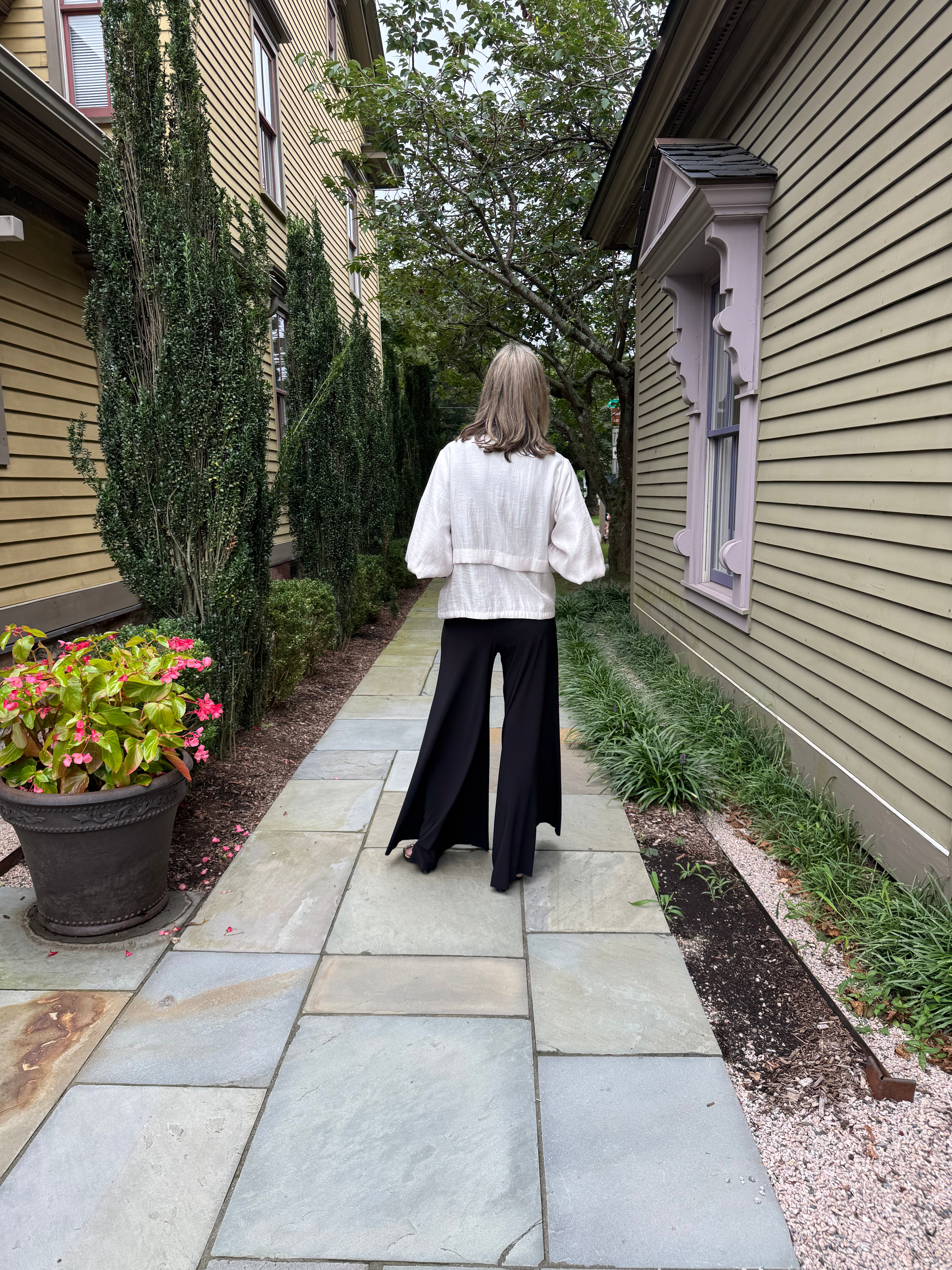 A woman viewed from behind walking on a stone path between houses, wearing a white puff-sleeve blouse and black wide-leg pants.