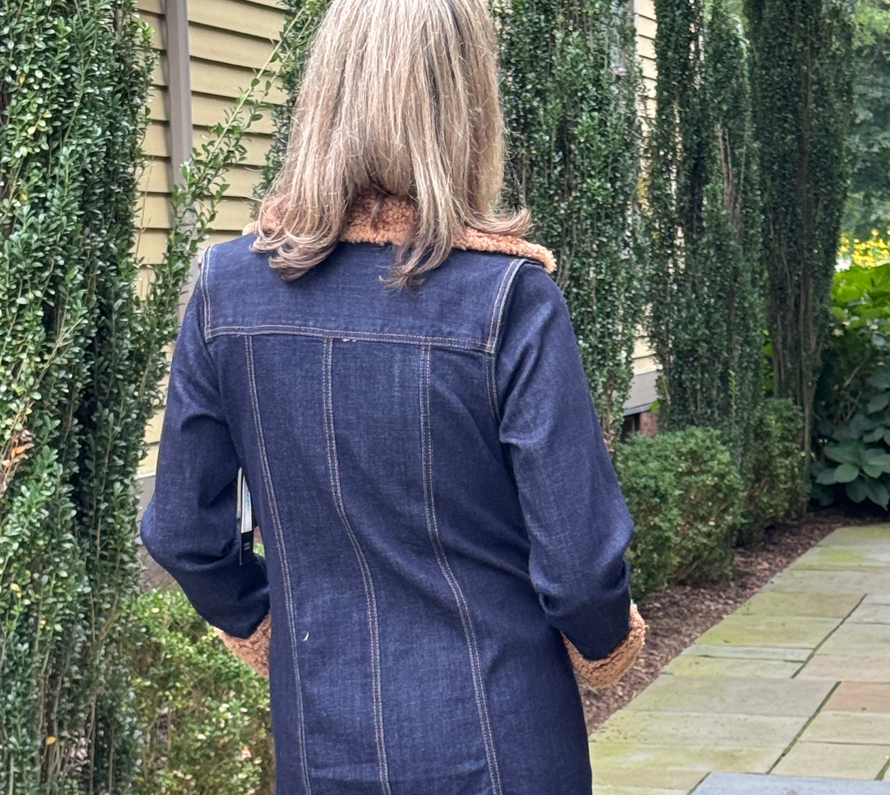 A woman viewed from behind wearing a dark blue denim coat with a brown faux-fur collar, standing on a stone patio in front of a house with green hedges.
