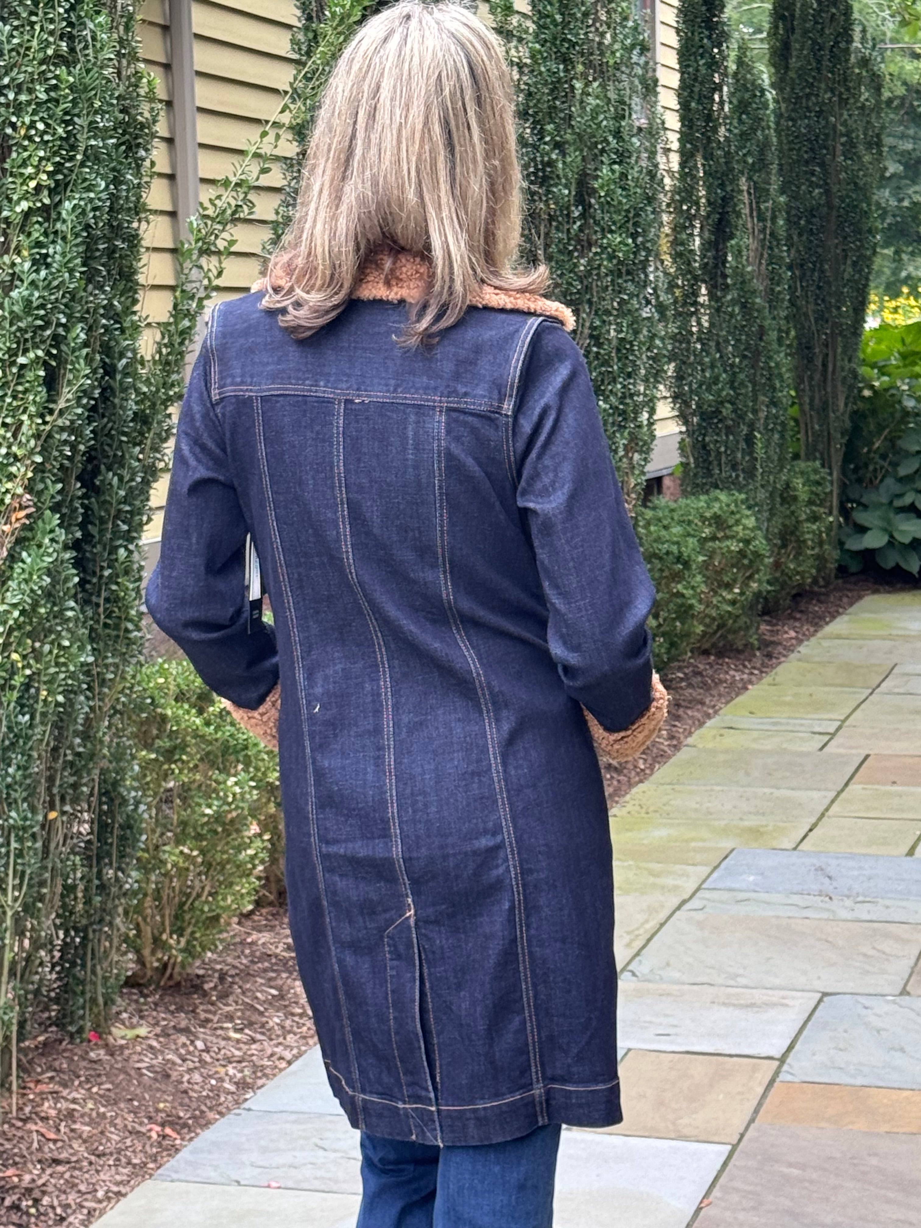 A woman viewed from behind wearing a dark blue denim coat with a brown faux-fur collar, standing on a stone patio in front of a house with green hedges.