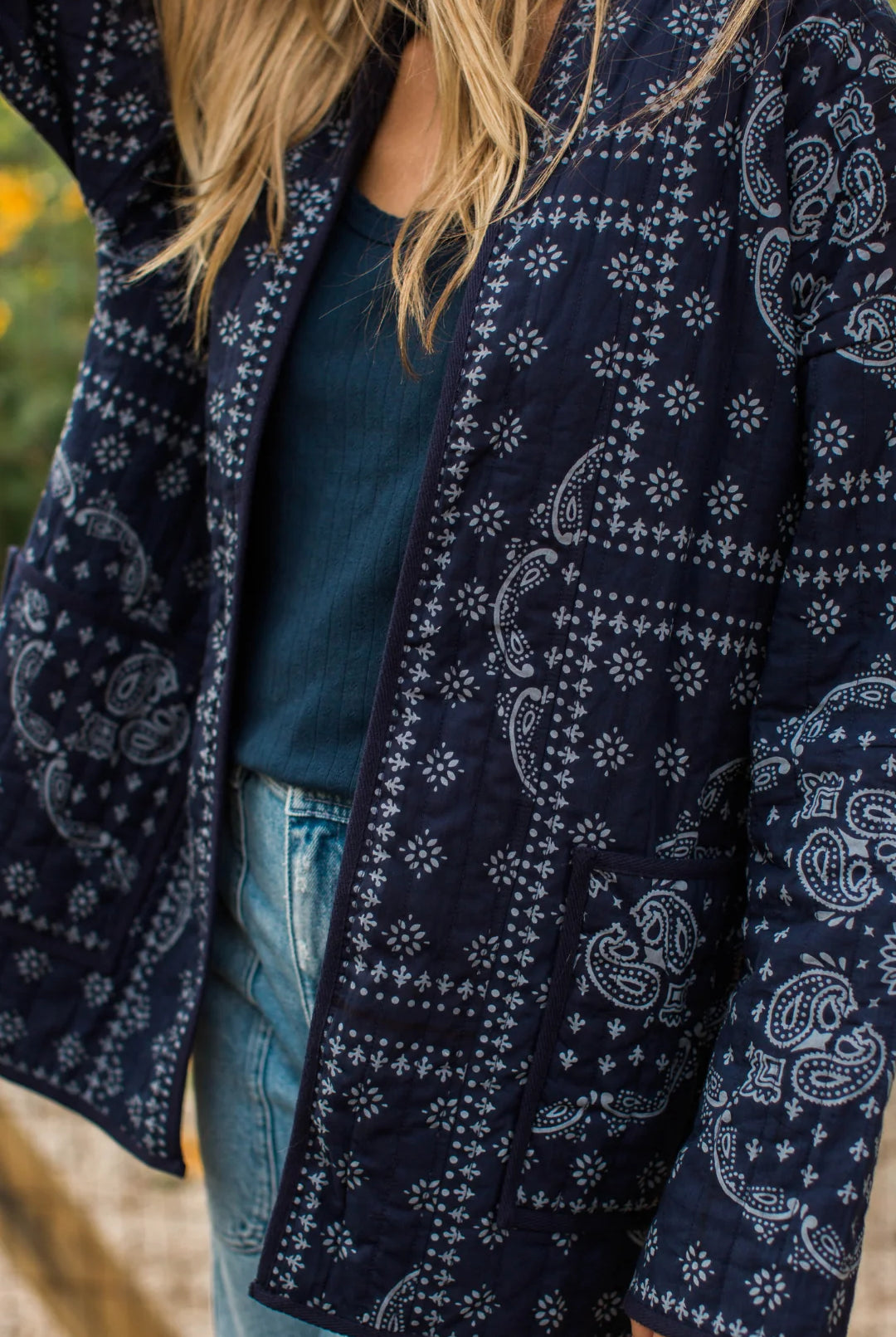 A close-up of a person wearing a navy blue quilted jacket with a white paisley and geometric bandana print, layered over a dark teal top and light blue jeans.