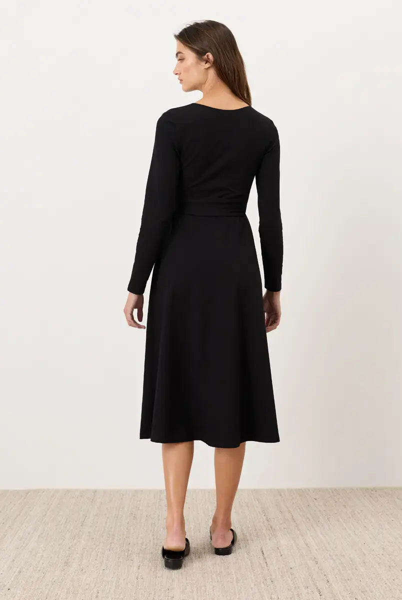 A woman wearing a black long-sleeve midi dress, shown from the back against a plain off-white wall and light wood floor.