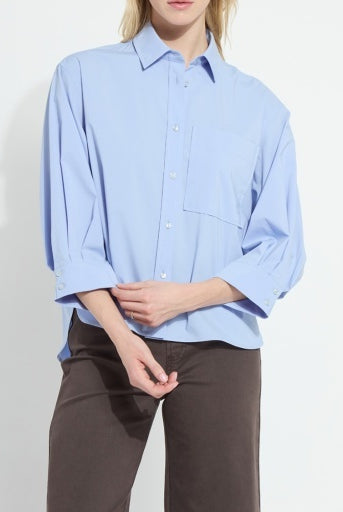 A model wearing a light blue button-down shirt with three-quarter sleeves and dark brown trousers, shown from the neck down against a plain white background.