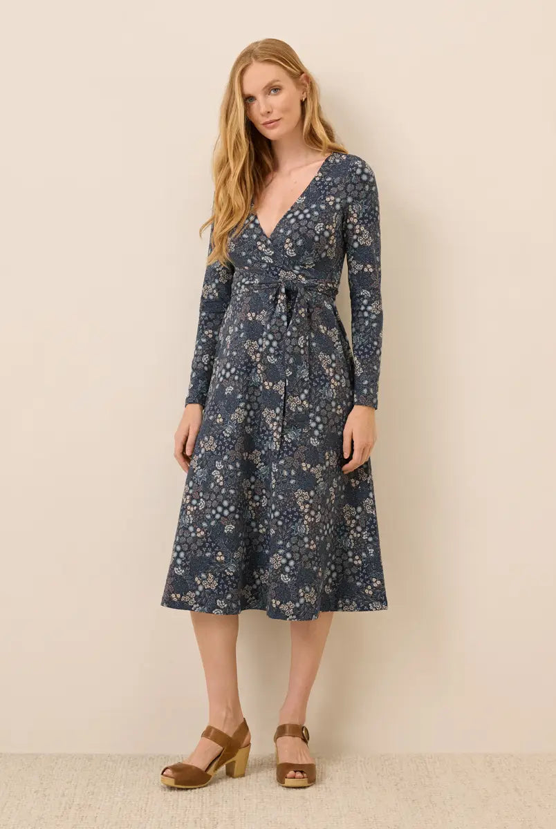 A woman modeling a long-sleeve wrap-style midi dress with a blue floral print and a matching tie belt, paired with brown heeled sandals, against a plain beige studio background.