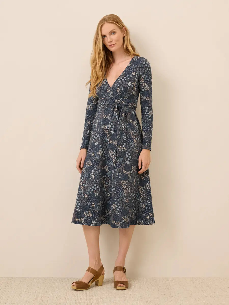 A woman modeling a long-sleeve wrap-style midi dress with a blue floral print and a matching tie belt, paired with brown heeled sandals, against a plain beige studio background.