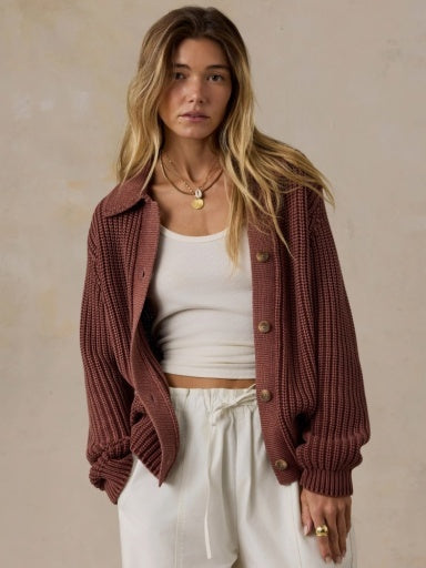 A woman wearing a rust-brown ribbed knit button-up cardigan over a white tank top and white drawstring pants, standing against a textured beige wall.