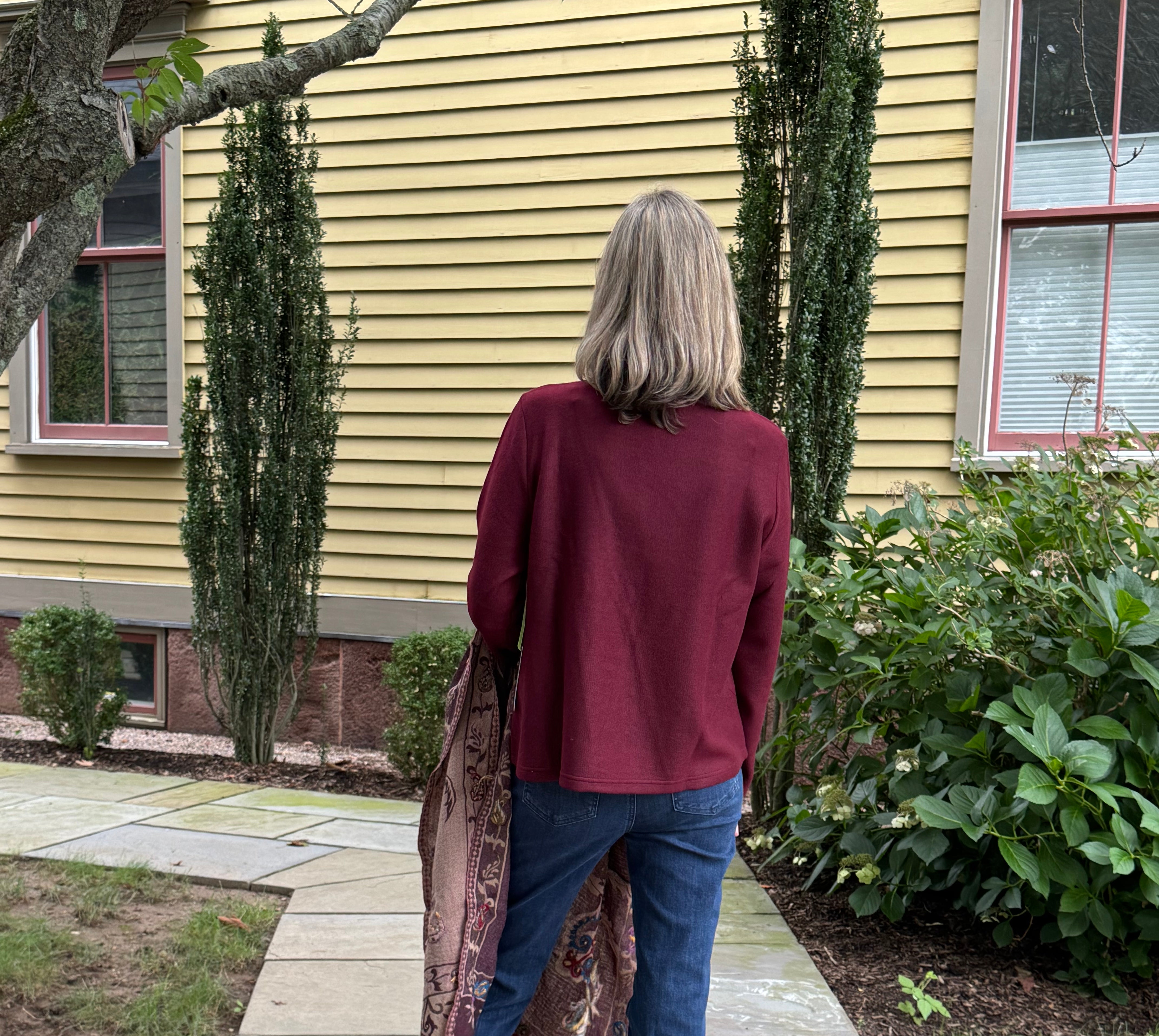 A woman viewed from behind stands on a stone walkway in front of a yellow house, wearing a burgundy long-sleeve top, blue jeans, and black sandals, while holding a patterned fringed scarf.