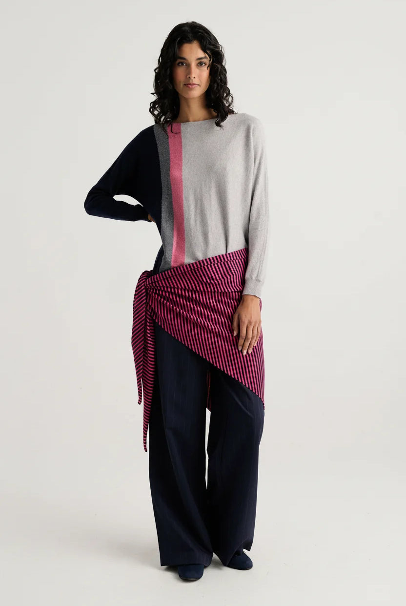 A model stands against a light gray studio background wearing a color-blocked knit sweater with navy, light gray, and pink sections, paired with dark wide-leg pants and a pink striped wrap tied around the waist.