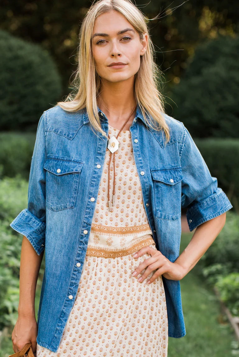 A woman standing outdoors in a garden wearing a light blue denim shirt over a cream patterned dress with a tan belt and a large white pendant necklace.