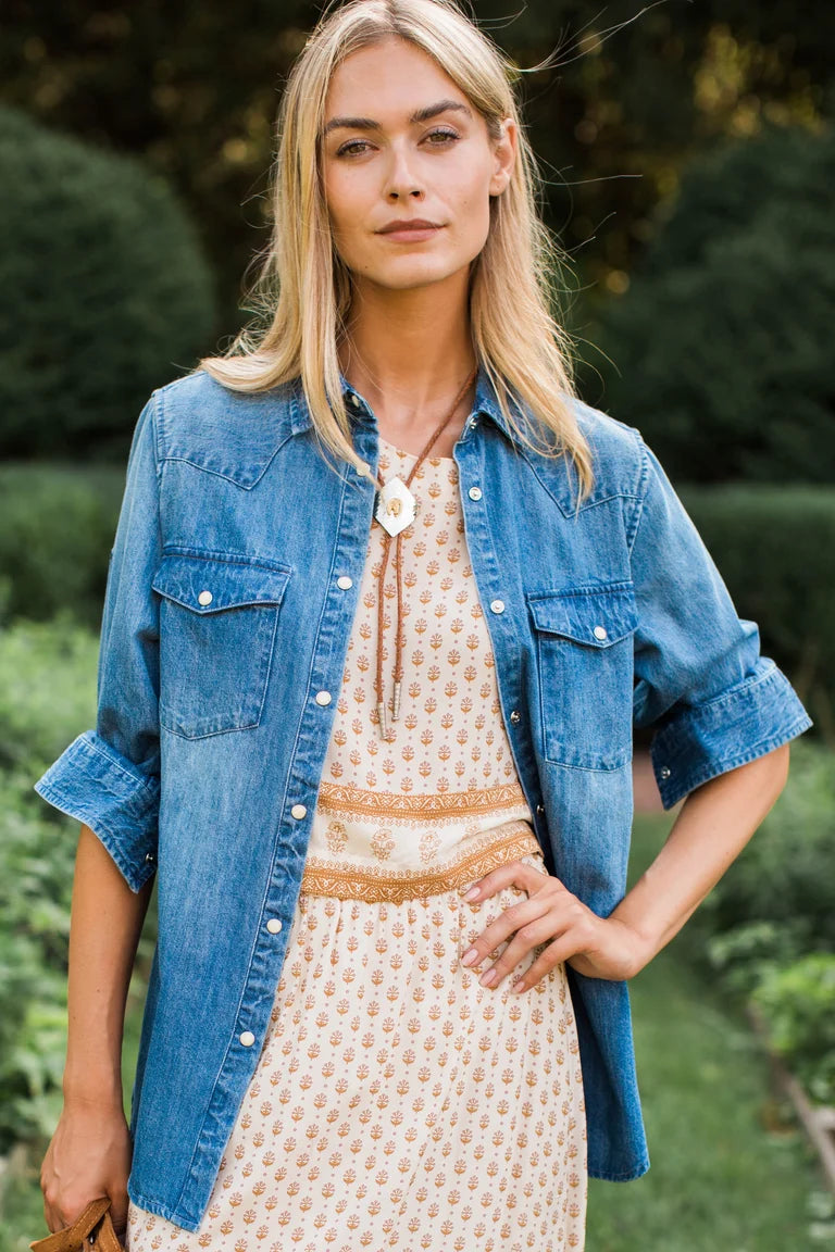 A woman standing outdoors in a garden wearing a light blue denim shirt over a cream patterned dress with a tan belt and a large white pendant necklace.