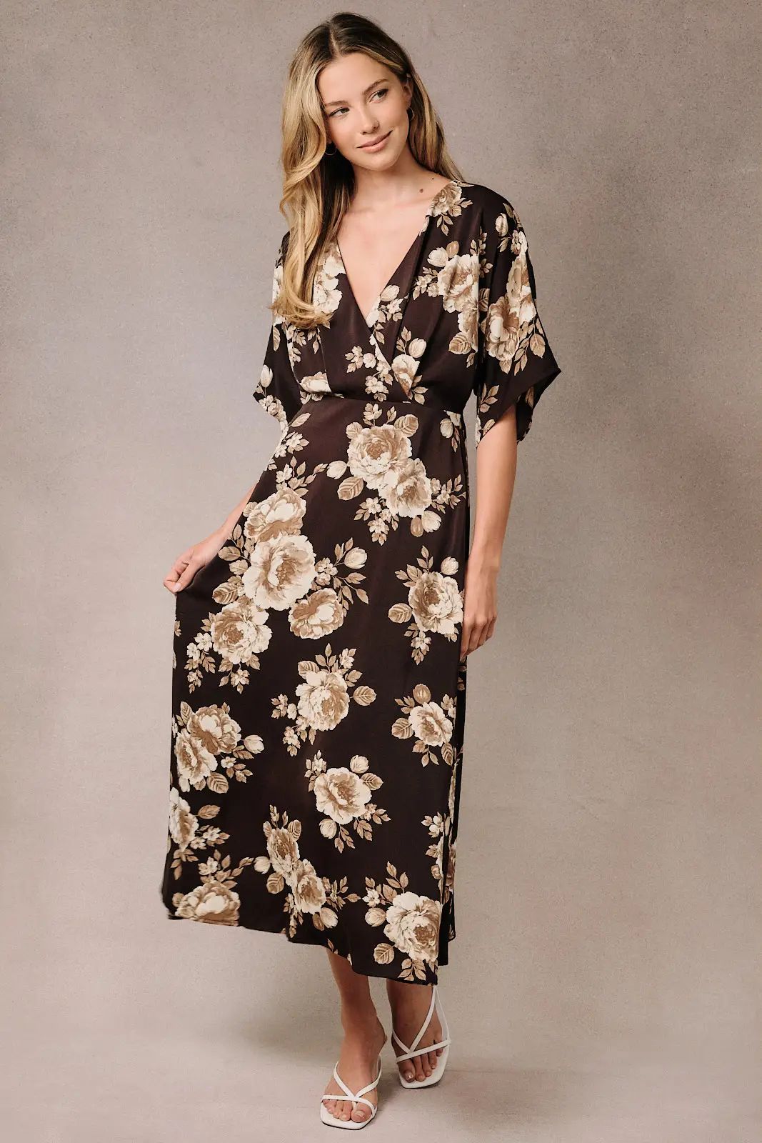 A woman modeling a dark brown midi dress with a beige and cream floral pattern, featuring a V-neckline, short flutter sleeves, and a matching fabric belt at the waist, set against a neutral studio backdrop.