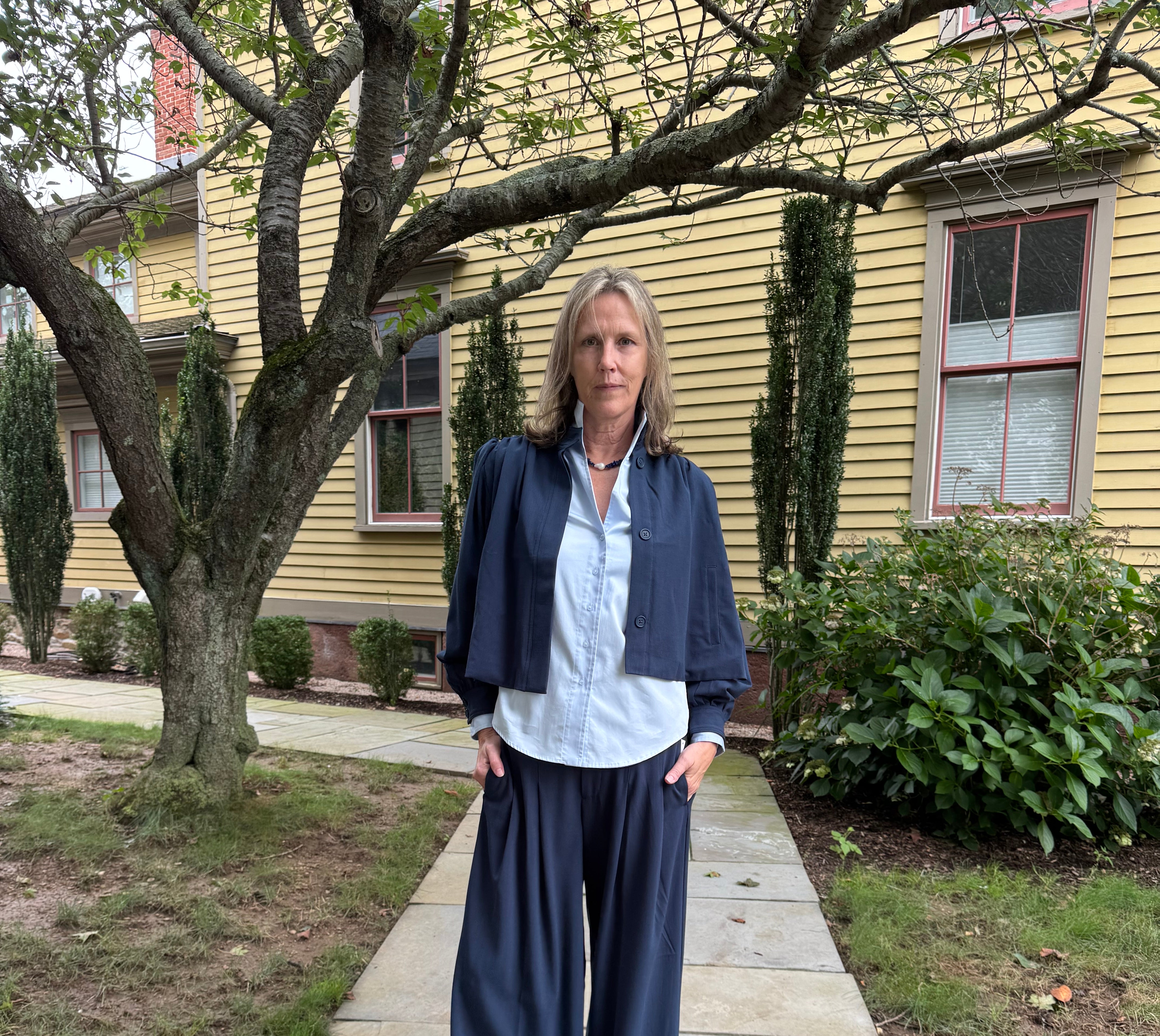 A woman stands on a stone walkway in front of a yellow house, wearing a navy blue cropped jacket, light blue button-up shirt, and navy wide-leg pants.
