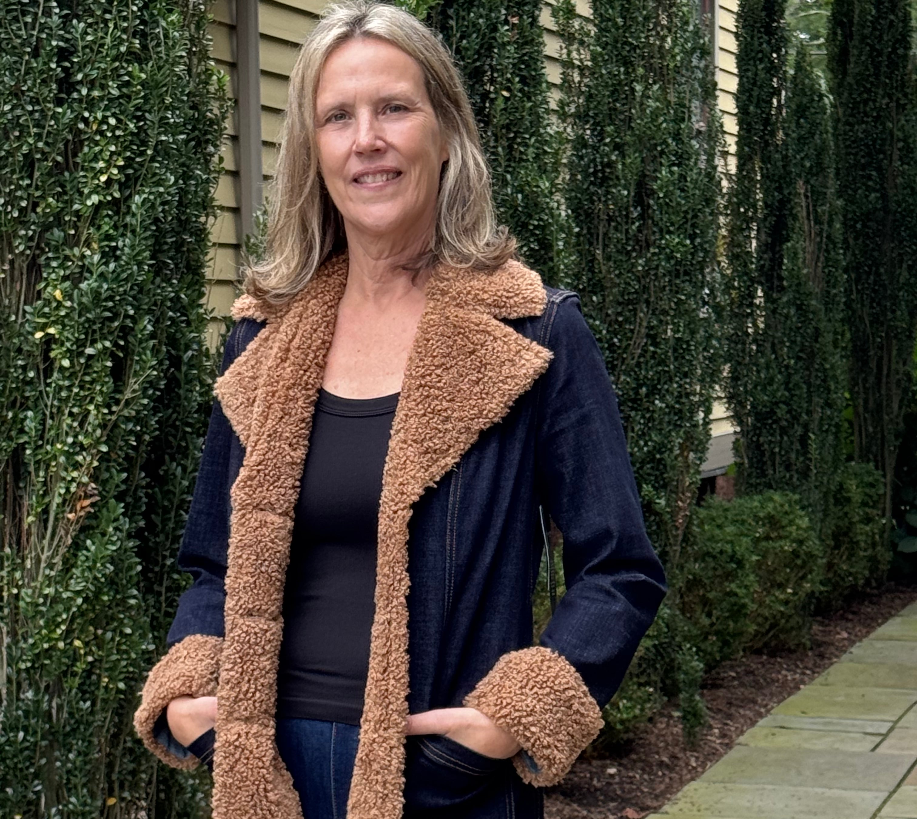 A woman wearing a dark blue denim-style coat with a thick tan sherpa lining, standing outdoors on a stone walkway with hedges and a house in the background.