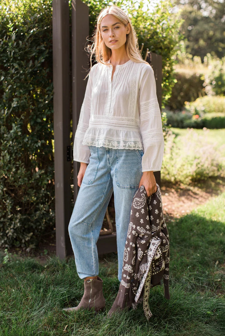 A woman stands outdoors on grass beside a wooden fence, wearing a white long-sleeve peplum blouse, light blue jeans, and brown ankle boots, while holding a dark paisley scarf.