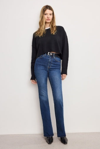 A woman modeling a black long-sleeve cropped sweater and high-waisted blue jeans with a black belt, standing against a plain off-white wall.