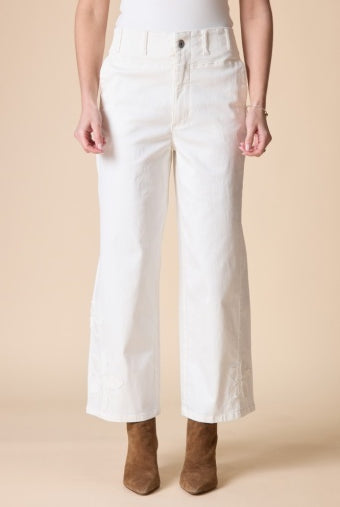 A model from the waist down wearing white cropped wide-leg jeans with tonal embroidery on the lower legs, paired with brown suede ankle boots against a solid beige background.