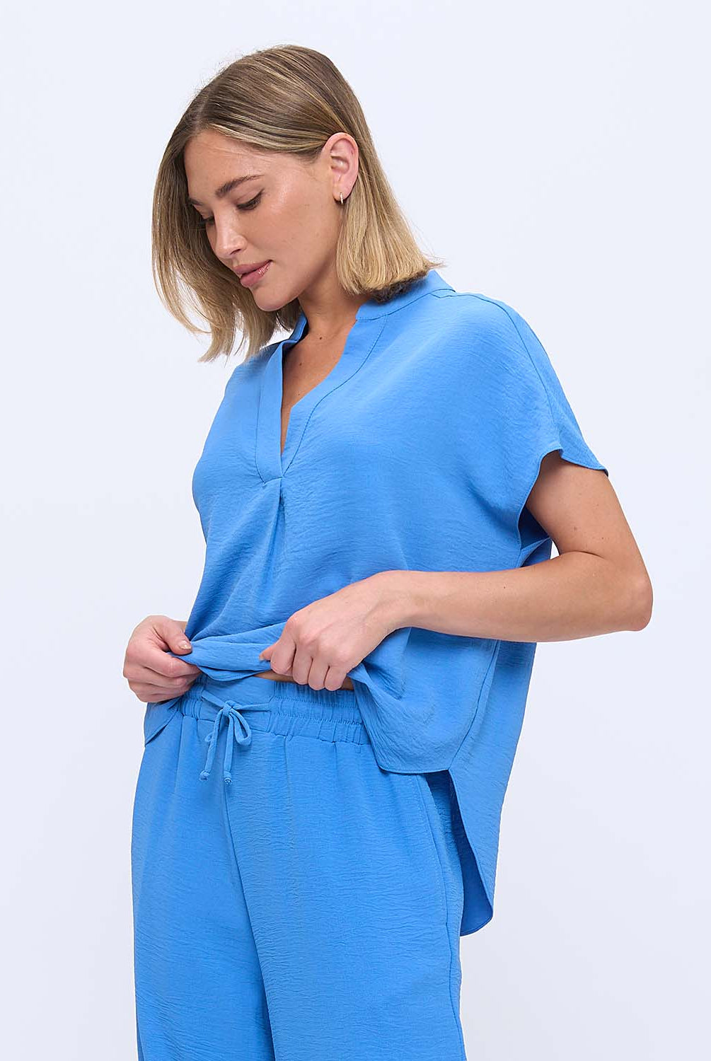 A woman wearing a bright blue short-sleeve top and matching drawstring pants, photographed against a clean light gray studio background.