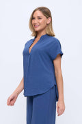 A woman modeling a blue short-sleeve scrub top and matching pants against a plain light gray studio background.