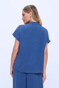 A woman shown from the back wearing a blue short-sleeve scrub top and matching blue pants against a light blue studio background.
