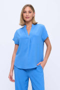 A woman modeling a bright blue scrub top and matching scrub pants against a plain light gray studio background.