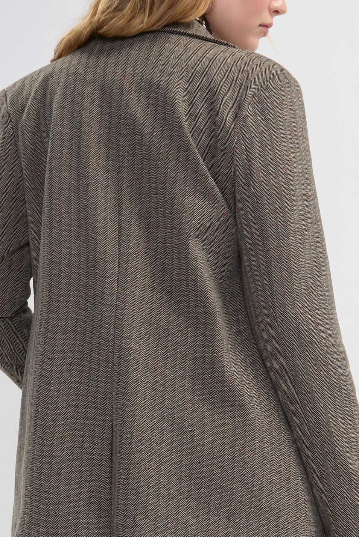 A woman shown from the back wearing a brown textured blazer with blue jeans, photographed against a light gray studio background.