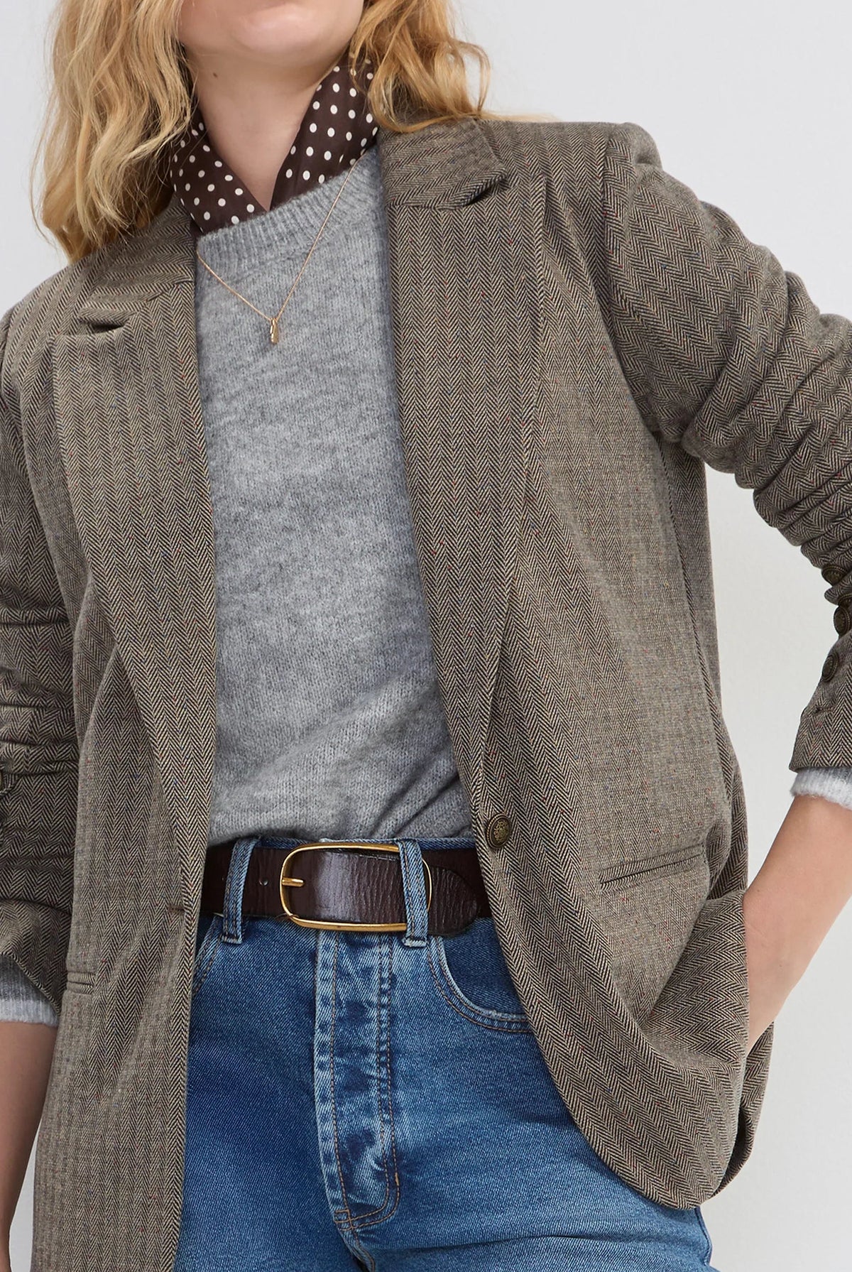 A woman modeling a brown and beige checkered blazer over a gray sweater, a polka-dot shirt collar, and blue jeans against a light gray studio background.