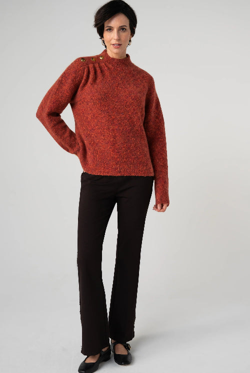 A woman modeling a textured rust-orange mock-neck knit sweater with decorative buttons on the shoulders, paired with black trousers and black flats, against a light gray studio background.