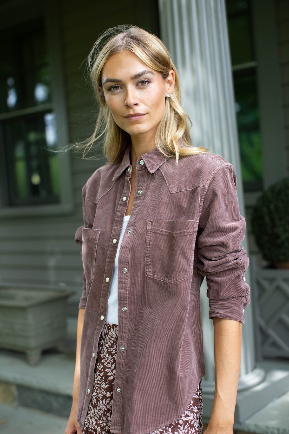 Woman standing on a porch in the MAREA western shirt in brown corduroy, worn open over a white tee and patterned skirt