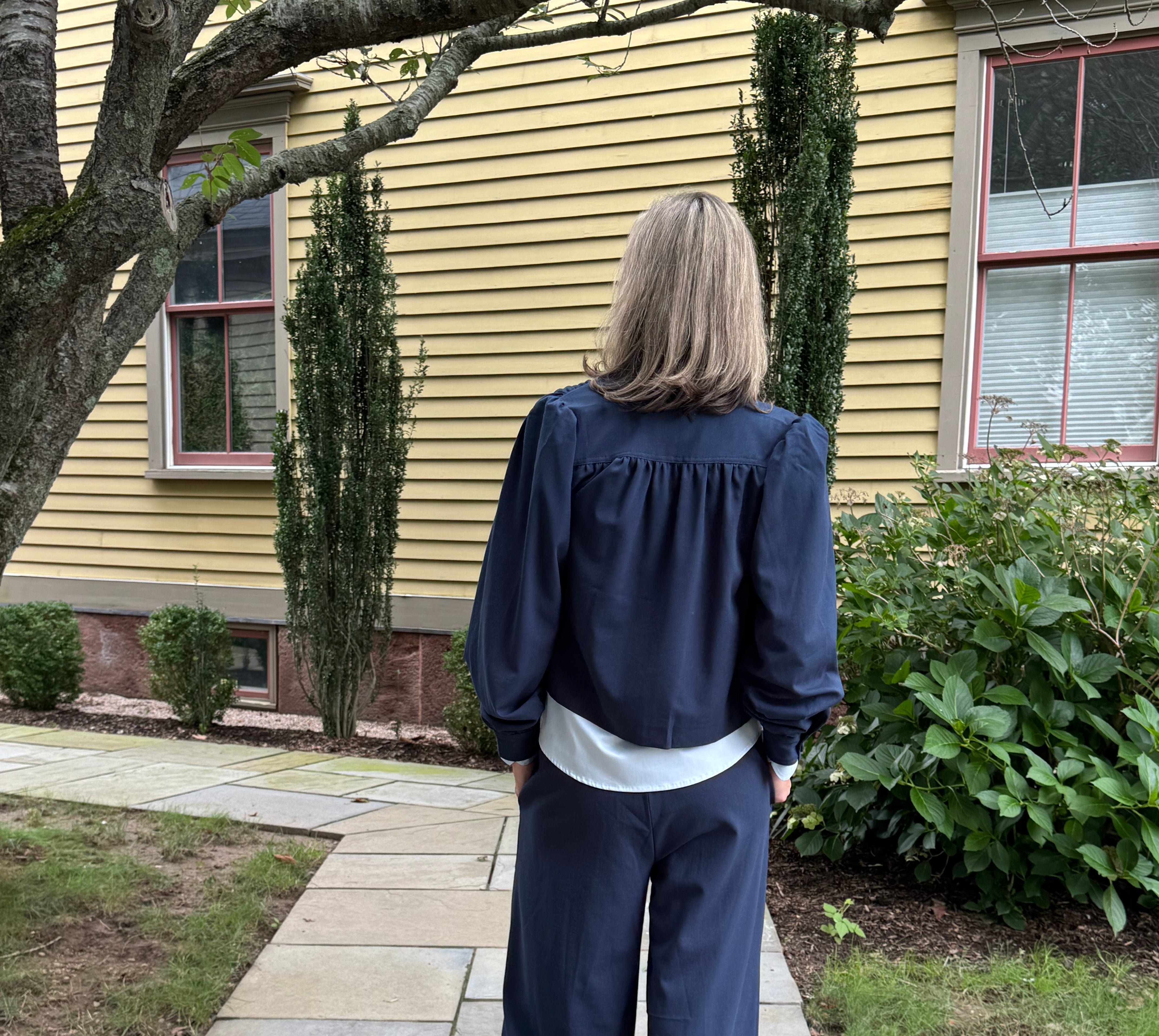 A woman stands on a stone walkway in front of a yellow house, viewed from behind. She is wearing a navy blue long-sleeve top with a gathered neckline and matching wide-leg pants.