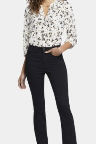 A woman modeling NYDJ High Rise Billie Mini Boot Petite bootcut jeans in Huntley black wash, paired with a floral print blouse and black heels, against a plain light studio background.