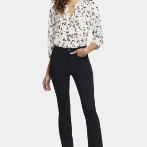 A woman modeling NYDJ High Rise Billie Mini Boot Petite bootcut jeans in Huntley black wash, paired with a floral print blouse and black heels, against a plain light studio background.
