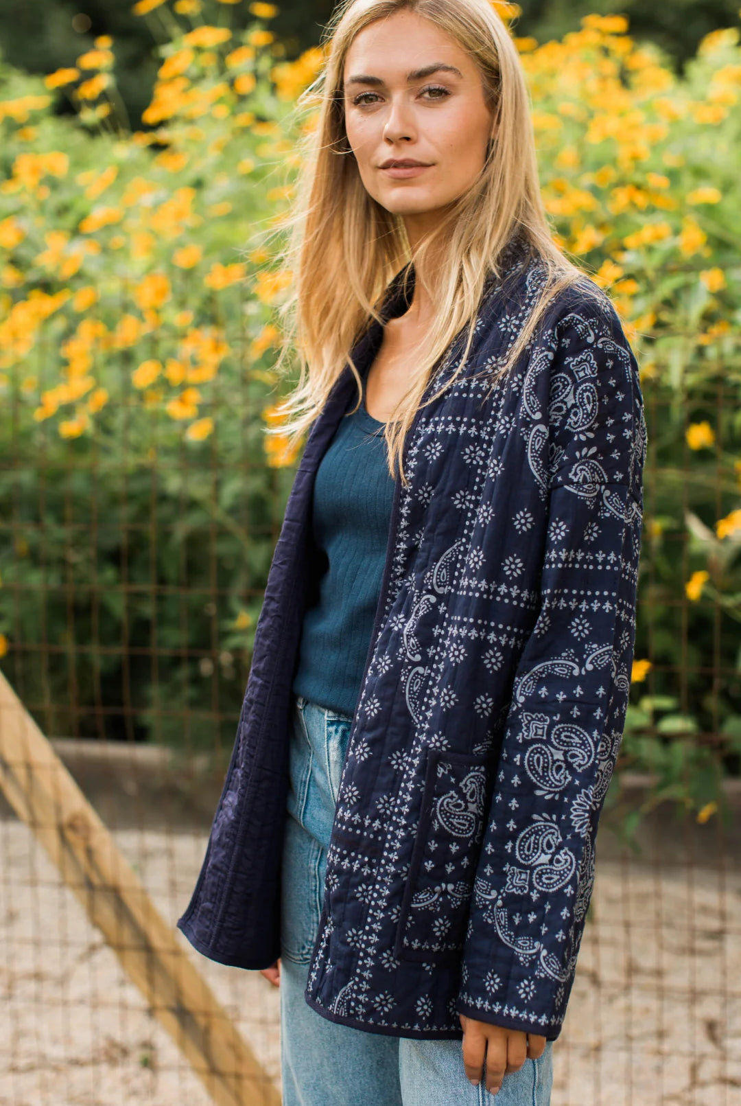 A woman standing outdoors in front of a garden of yellow flowers, wearing a navy blue quilted jacket with a white paisley bandana print, a dark teal top, and light blue jeans.