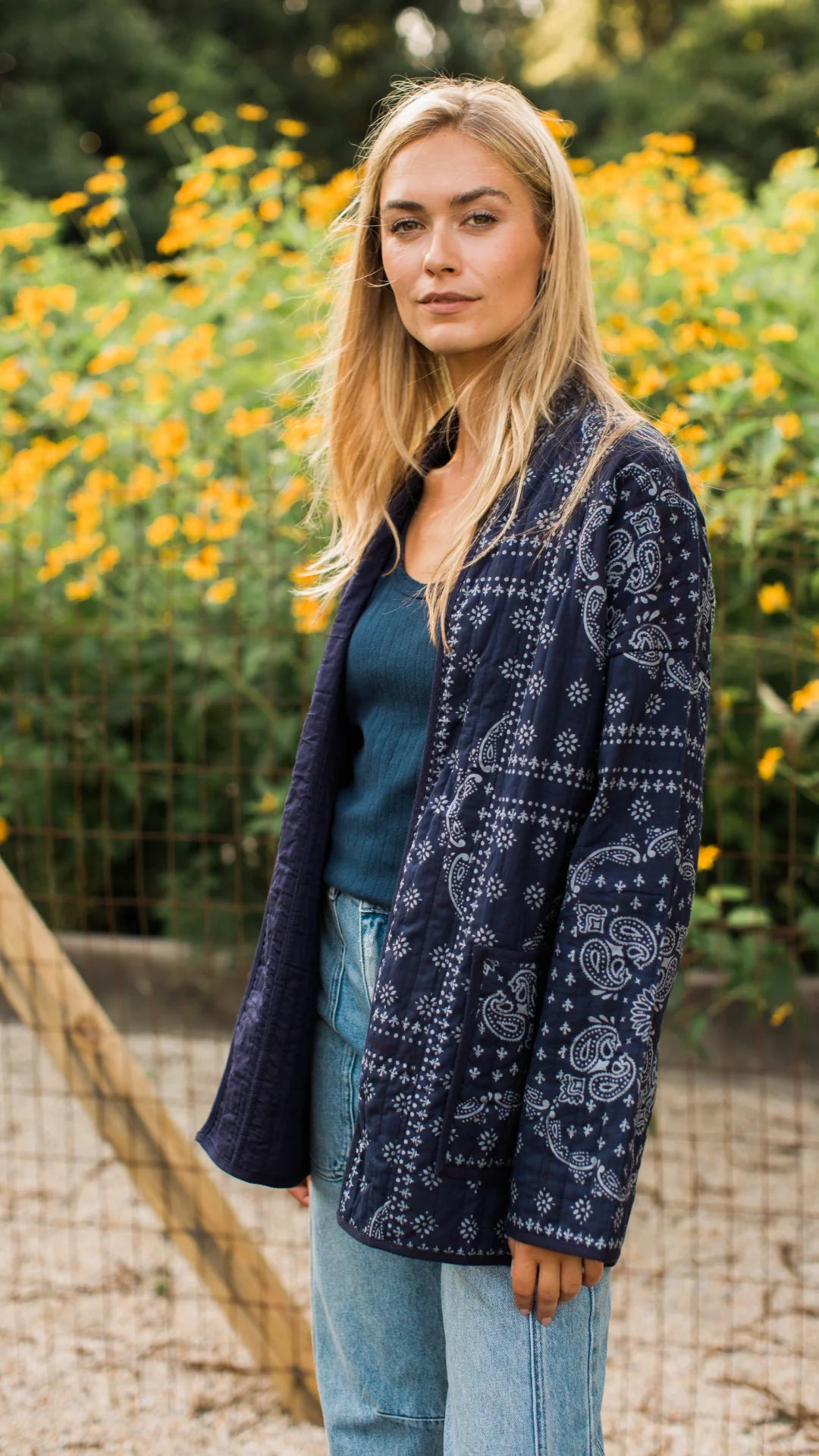 A woman standing outdoors in front of a garden of yellow flowers, wearing a navy blue quilted jacket with a white paisley bandana print, a dark teal top, and light blue jeans.